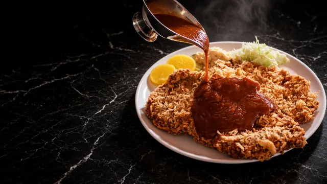 King Katsu: a wide pork cutlet on a white plate, sauce being poured, on black marble