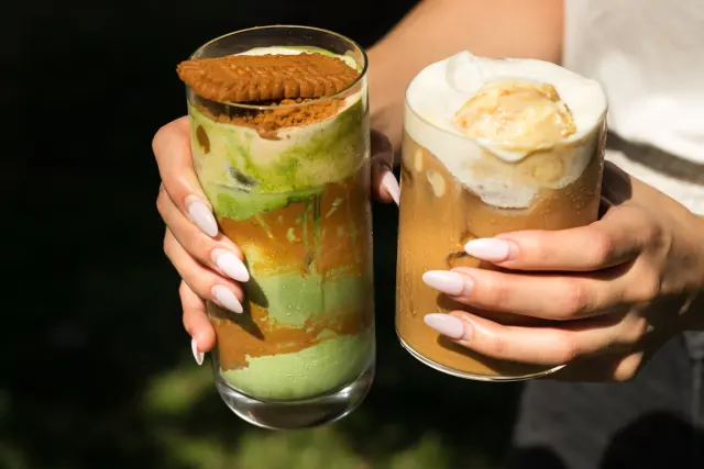 Two Cafe Mallow iced drinks held up in daylight, one matcha with a biscuit on top, one iced coffee with cream